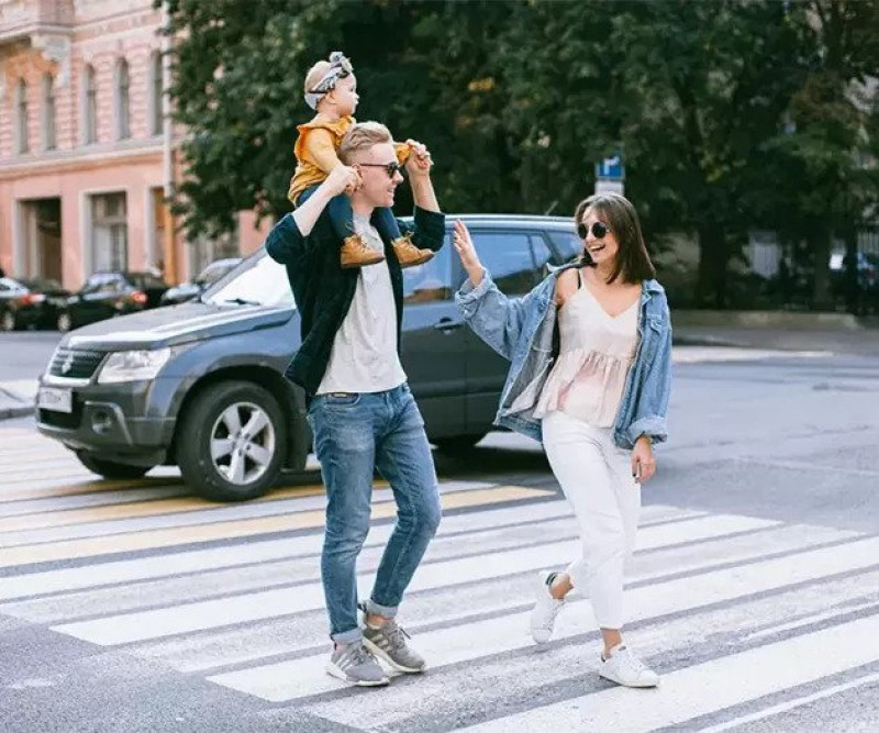 Nasmejana družina prečka cesto na prehodu za pešce oče nosi otroka na ramenih mati pa mu daje petko medtem ko hodita v sproščenem mestnem okolju