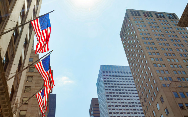 Ameriške zastave visijo ob strani visoke stavbe v New Yorku medtem ko sonce sije nad mestnimi nebotičniki pod jasnim nebom