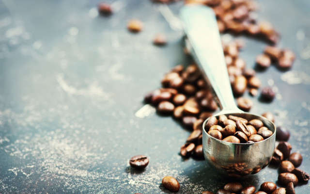 Kovinska žlica je napolnjena s praženimi kavnimi zrni ki so raztresena po temni površini