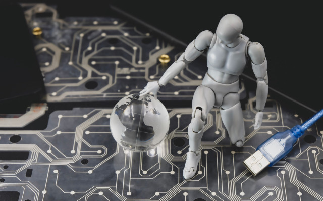 Srebrna robotska figura na vezju kleči ob prosojni krogli z zemljevidom sveta poleg modrega USB kabla