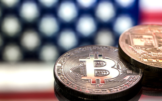 Dva kovanca z bitcoin simbolom ležita na odsevni površini pred zamegljenim ozadjem ameriške zastave
