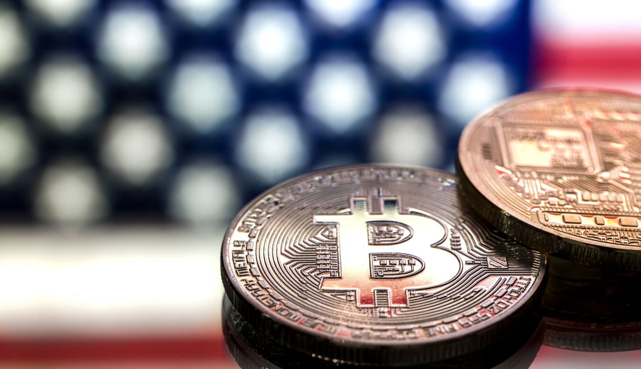 Dva kovanca z bitcoin simbolom ležita na odsevni površini pred zamegljenim ozadjem ameriške zastave