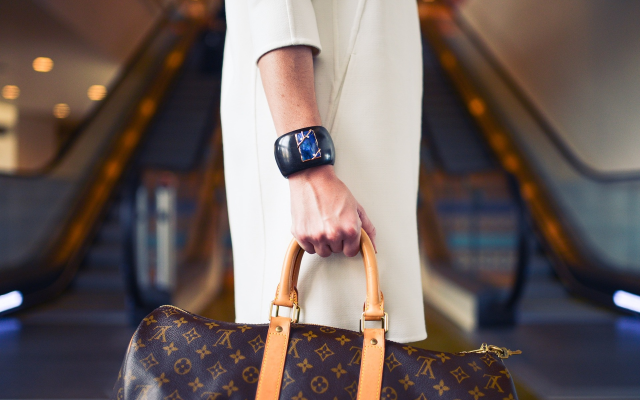 Ženska v beli obleki stoji pred tekočimi stopnicami in v roki drži luksuzno torbo Louis Vuitton