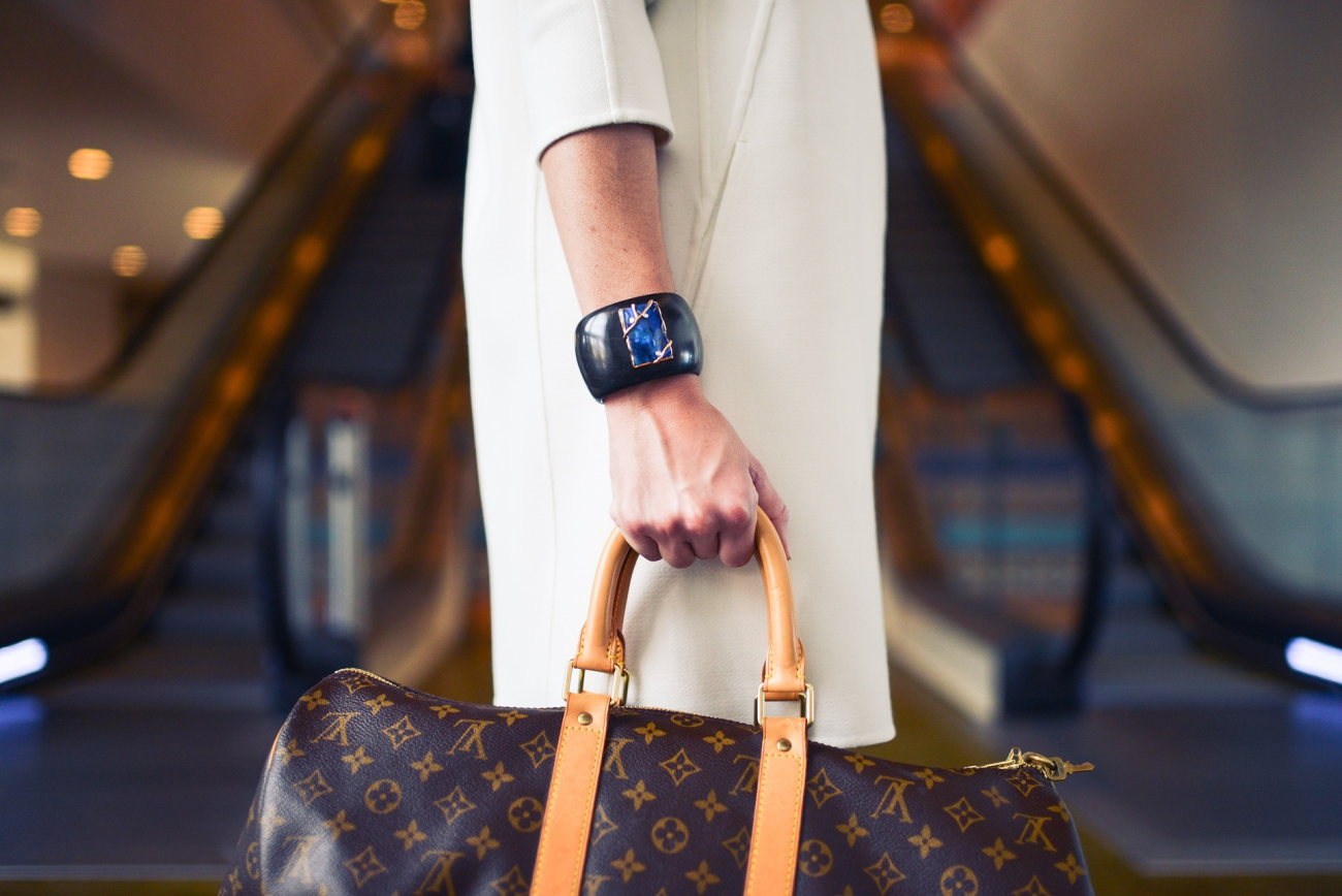 Ženska v beli obleki stoji pred tekočimi stopnicami in v roki drži luksuzno torbo Louis Vuitton