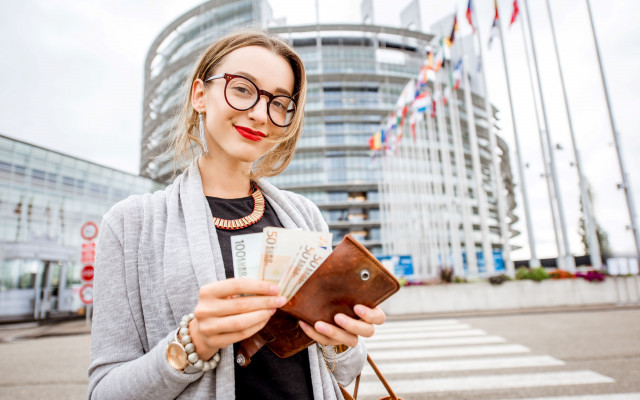 Ženska z očali in rdečo šminko stoji pred Evropskim parlamentom in drži evrske bankovce v denarnici