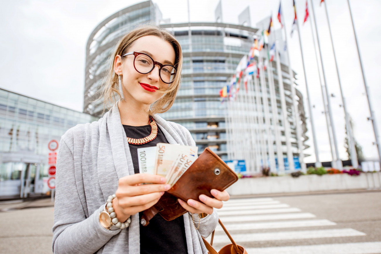 Ženska z očali in rdečo šminko stoji pred Evropskim parlamentom in drži evrske bankovce v denarnici