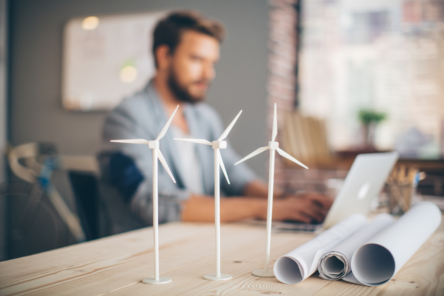 Na sliki je moški, ki dela za računalnikom, medtem ko so v ospredju makete vetrnih turbin in arhitekturni načrti kar simbolizira delo na področju obnovljivih virov energije.