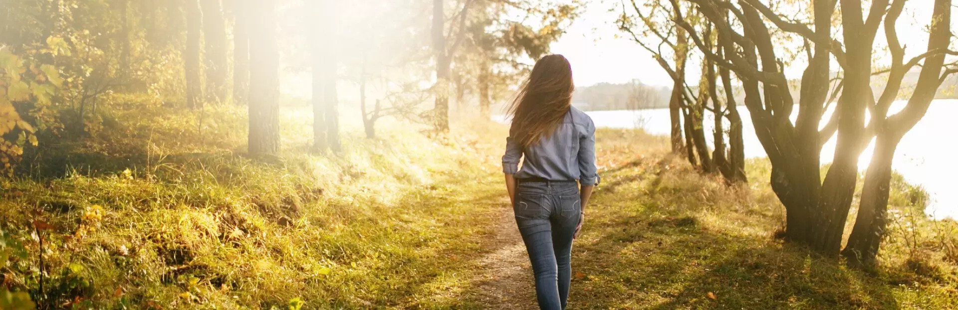 Ženska hodi po gozdni poti ob jezeru v toplem sončnem popoldnevu obdana z jesensko obarvanim drevjem in mirno naravo