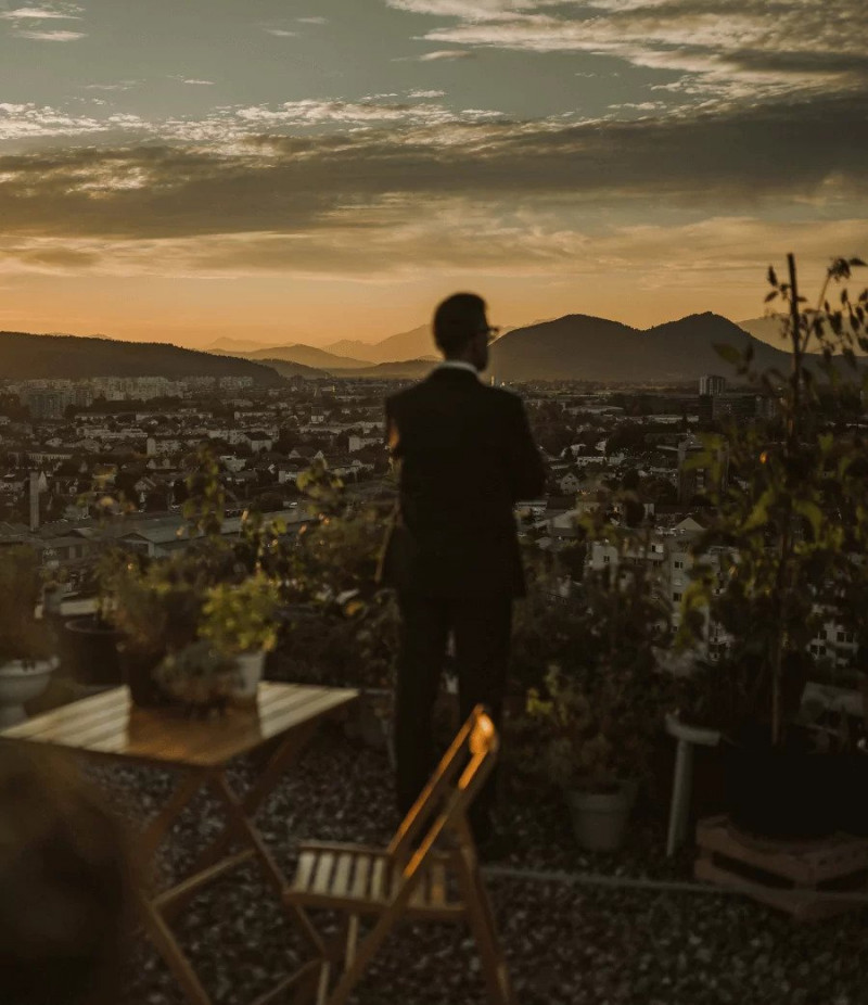 Moški v obleki stoji na terasi s pogledom na mesto ob sončnem zahodu obdan z rastlinami in vrtnim pohištvom ter zre v daljavo nad mestno pokrajino