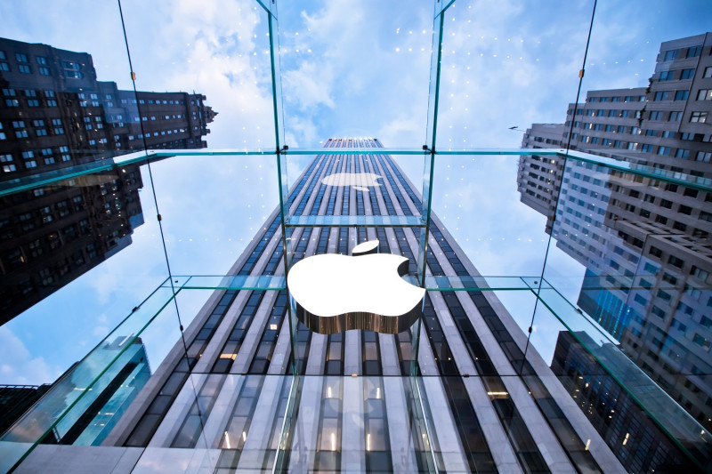Pogled navzgor skozi stekleno streho razkriva velik bel logotip podjetja Apple in visok poslovni nebotičnik med stolpnicami v mestu
