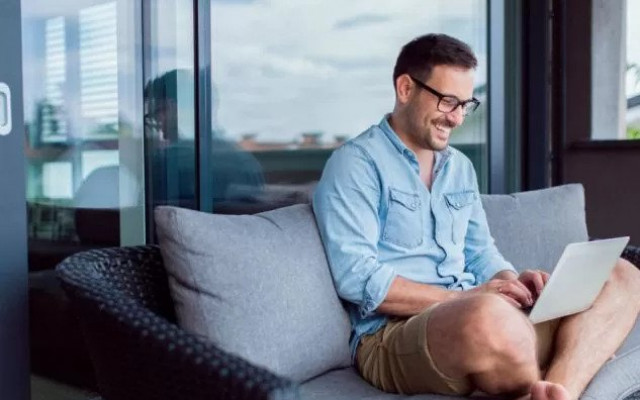 Nasmejan moški v kratkih hlačah in srajci sedi sproščeno na terasi z računalnikom v naročju in dela ali brska po spletu medtem ko je pametni telefon položen ob njem