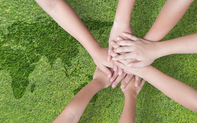 Na sliki so združene roke več oseb na travi kar simbolizira enotnost sodelovanje in skupnost.