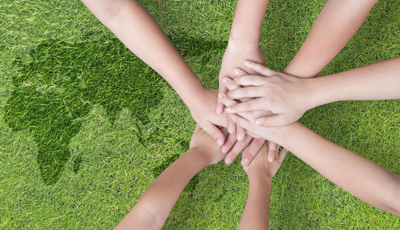 Na sliki so združene roke več oseb na travi kar simbolizira enotnost sodelovanje in skupnost.