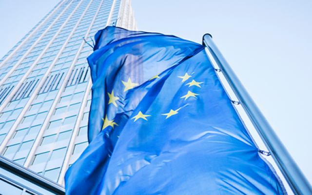 Na sliki je zastava Evropske unije ki plapola pred visoko sodobno stekleno stolpnico