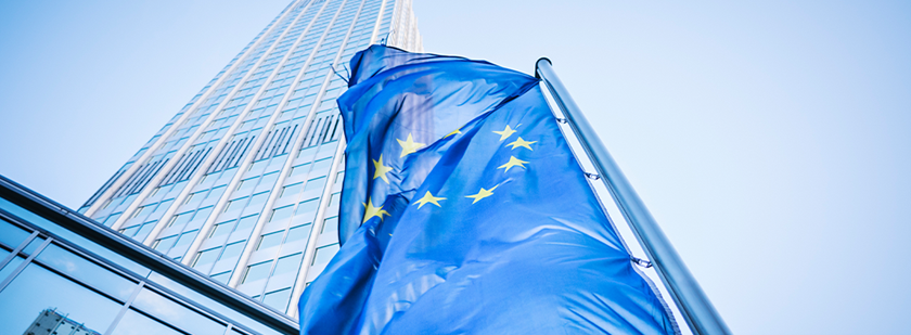 Na sliki je zastava Evropske unije ki plapola pred visoko sodobno stekleno stolpnico