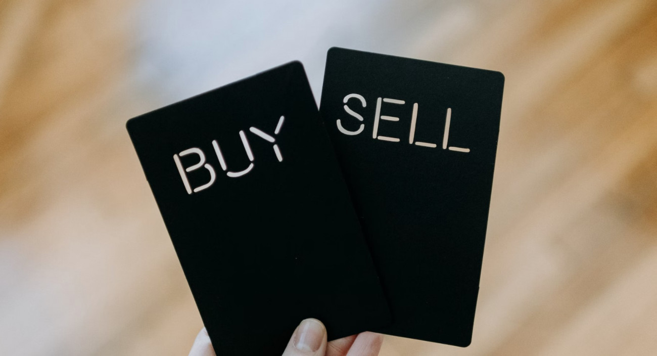 Roka drži dve črni kartici z izrezanima napisoma buy in sell kar simbolizira odločanje med nakupom in prodajo