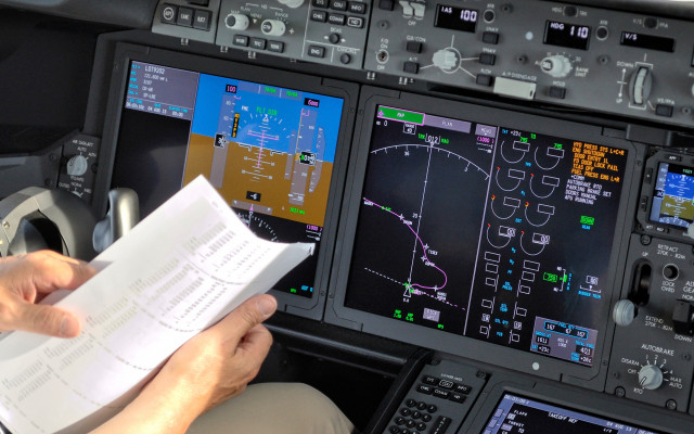 Pilot v pilotski kabini drži navigacijski seznam pred sodobnimi zasloni z letalskimi instrumenti in letalsko potjo