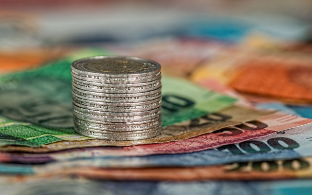 Na sliki je kup kovancev ki stoji na kupu barvitih bankovcev kar simbolizira bogastvo finance ali varčevanje
