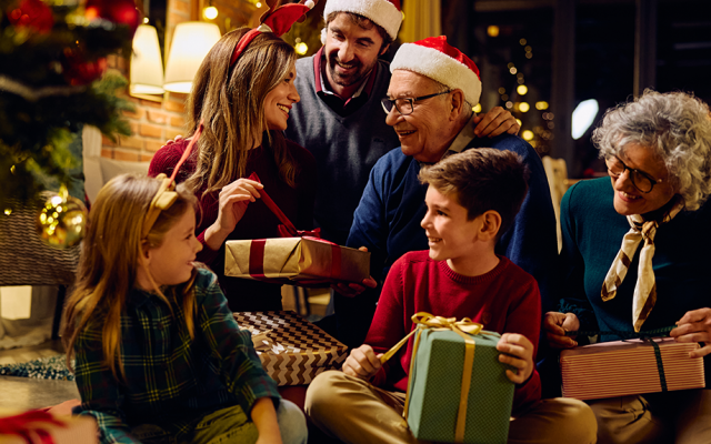 Družina v prazničnem vzdušju sedi ob božičnem drevesu in si z nasmehi izmenjuje darila