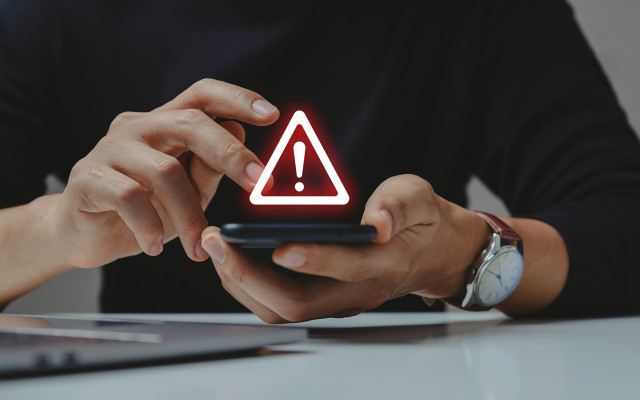 Oseba drži pametni telefon z opozorilnim znakom z rdečim klicajem na zaslonu kar nakazuje na varnostno ali sistemsko težavo