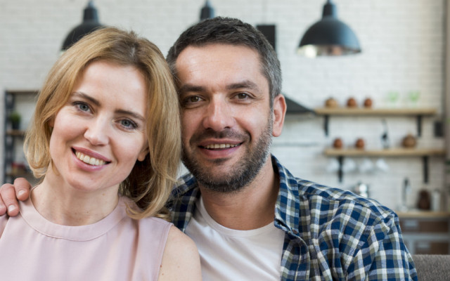 Nasmejan par pozira skupaj na kavču v svetli sodobni kuhinji z belim zidom in visečimi lučmi v ozadju
