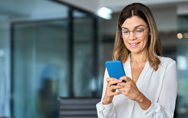 Slika poslovno oblečene ženske v sodobni pisarni, ki gleda v mobilni telefon.