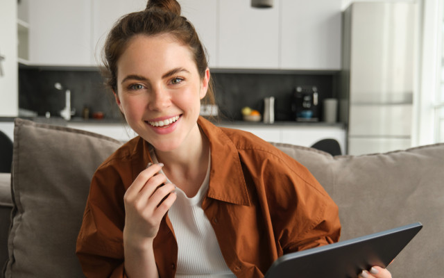 Zadovoljna ženska sedi na kavču, v roki ima tablični računalnik.