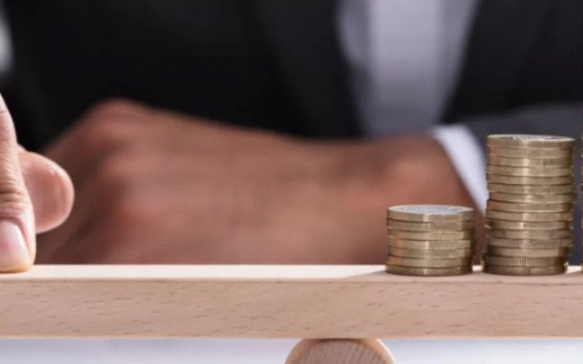 Moški v poslovni obleki s prstom drži ravnotežje na leseni deski ki stoji na sredinskem valju z naraščajočimi kupčki kovancev na desni strani kar simbolizira uravnoteženje financ ali naložb