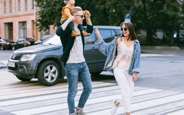 Nasmejana družina prečka cesto na prehodu za pešce oče nosi otroka na ramenih mati pa mu daje petko medtem ko hodita v sproščenem mestnem okolju