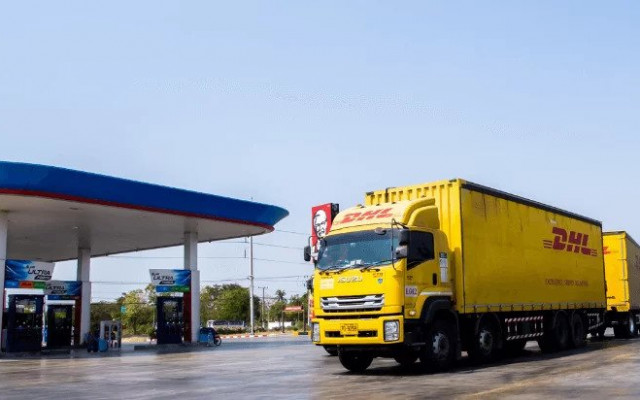 Rumeni DHL tovornjak stoji na velikem parkirišču ob bencinski črpalki z modro rdečo streho na sončen dan v bližini mesta 