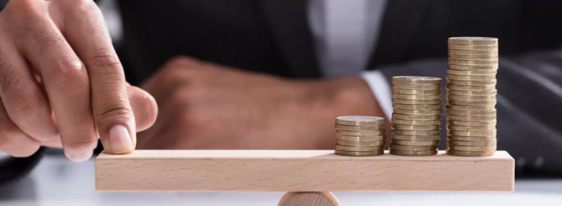 Moški v poslovni obleki s prstom drži ravnotežje na leseni deski ki stoji na sredinskem valju z naraščajočimi kupčki kovancev na desni strani kar simbolizira uravnoteženje financ ali naložb