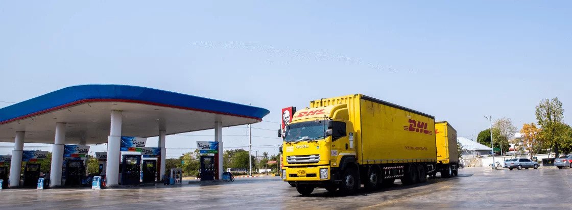 Rumeni DHL tovornjak stoji na velikem parkirišču ob bencinski črpalki z modro rdečo streho na sončen dan v bližini mesta 