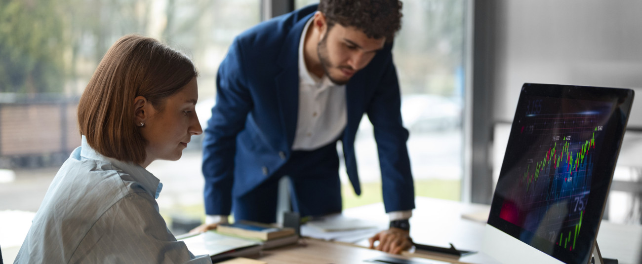 Slika prikazuje finančna strokovnjaka, žensko in moškega, ki gledata na računalniški zaslon s finančno analizo.