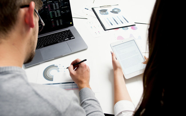 osebi analizirata podatke pri mizi; eden piše po natisnjenih grafih ob odprtem prenosniku, drugi bere dokument na tablici