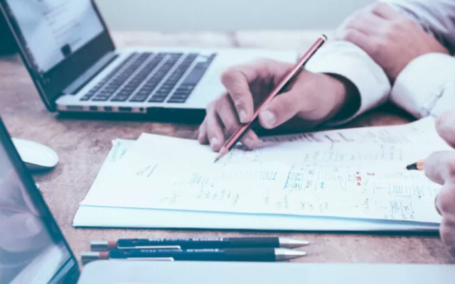 Slika prikazuje roko osebe, ki s svinčnikom kaže na podatek, zapisan na listu papirja