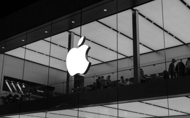 Črno bela fotografija trgovine Apple z velikim svetlečim logotipom in ljudmi ki sedijo za mizami 