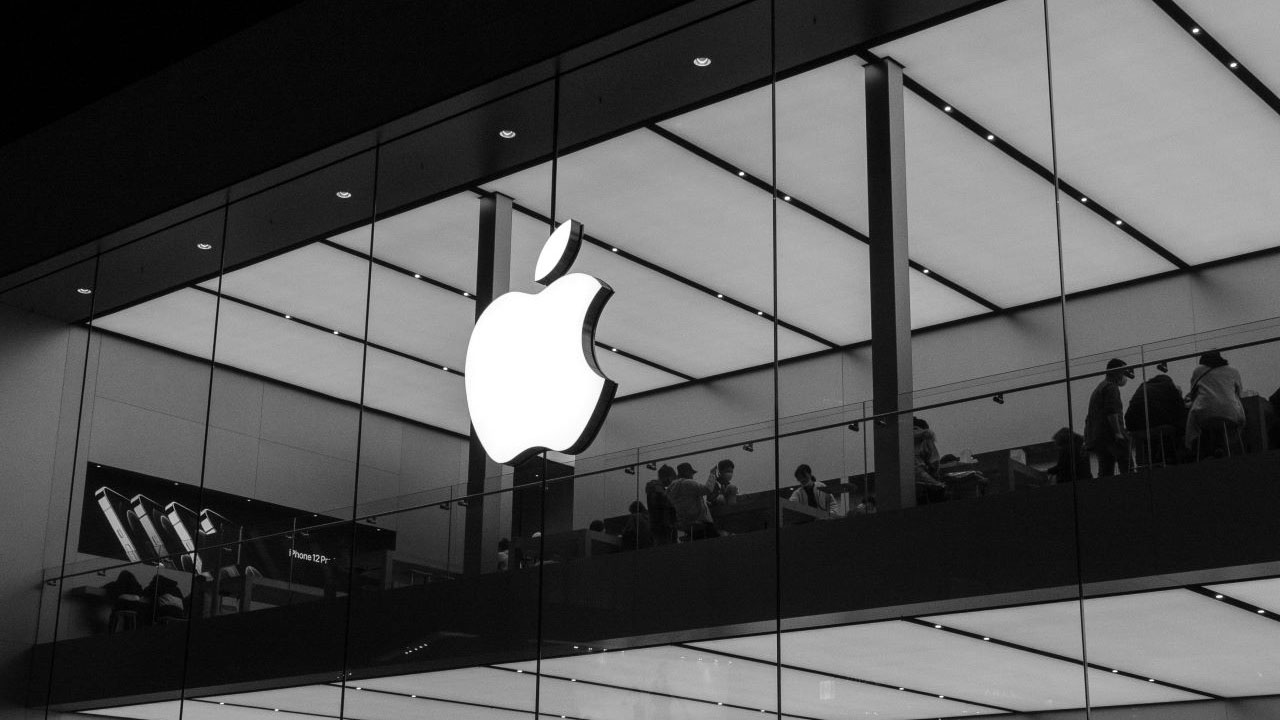 Črno bela fotografija trgovine Apple z velikim svetlečim logotipom in ljudmi ki sedijo za mizami 