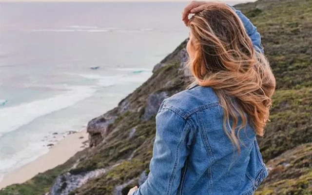 Ženska v jeans jakni stoji na klifu in gleda proti oceanu z vetrom v laseh
