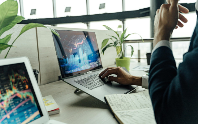 Oseba dela na prenosniku z izpisanimi finančnimi grafikoni, na mizi pa so zapiski in rastlina