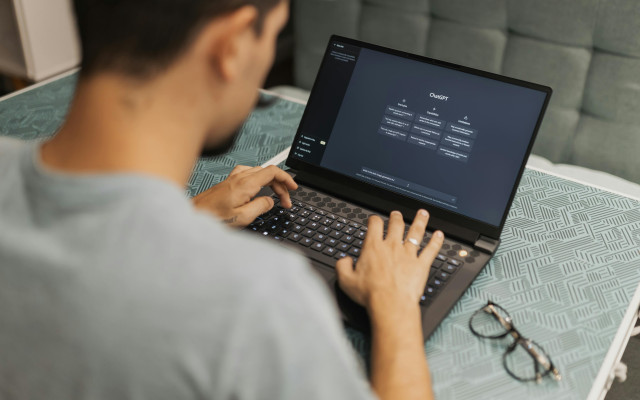 Oseba sedi za prenosnikom in tipka, medtem ko je na zaslonu odprt vmesnik ChatGPT; ob računalniku ležijo očala