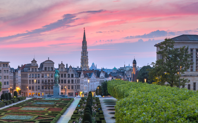Panoramski pogled na Bruselj ob sončnem zahodu z urejenim parkom v ospredju in mestnimi stavbami v ozadju.