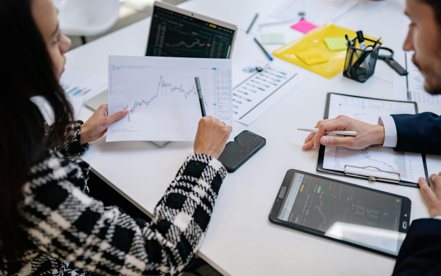 Osebi ob mizi analizirata finančne grafe na papirju, prenosniku in tablici