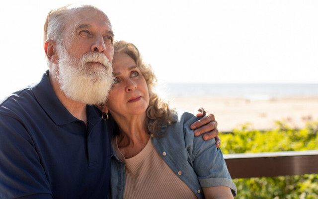 Starejši par sedi na klopi ob plaži in se objema medtem ko gleda v daljavo