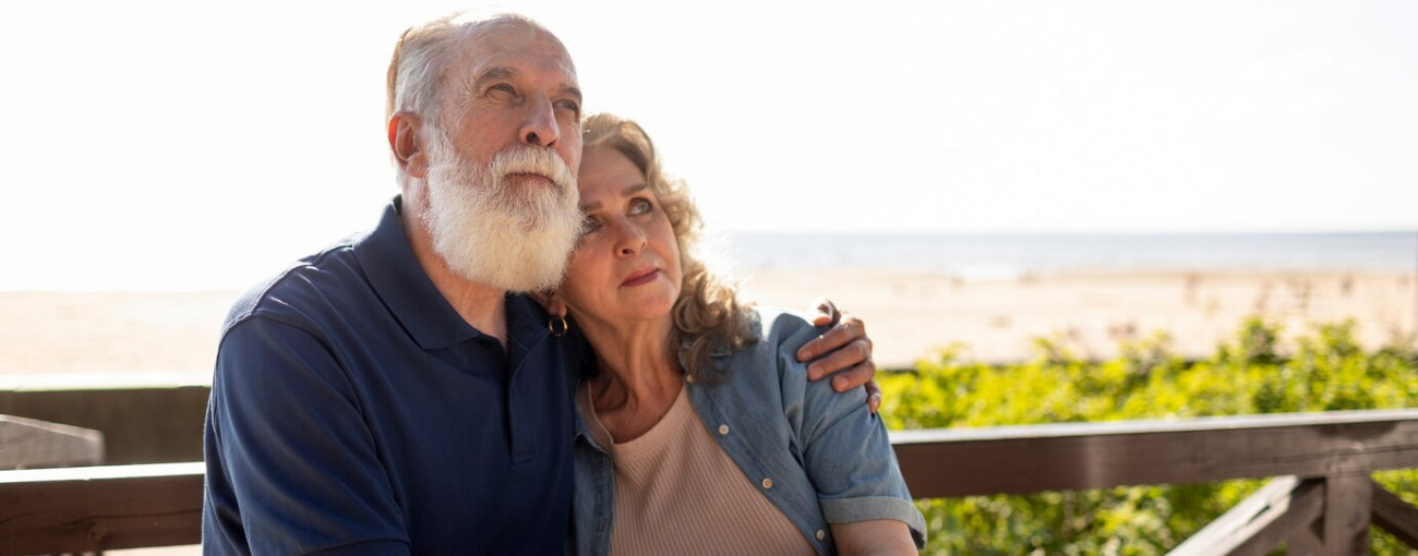 Starejši par sedi na klopi ob plaži in se objema medtem ko gleda v daljavo