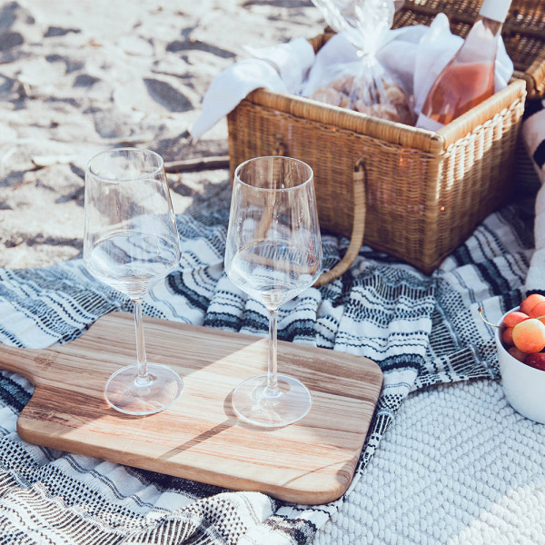 Na piknik odeji na plaži stojita prazna vinska kozarca ob leseni deski poleg košare s hrano steklenico vina in skodelico jagod