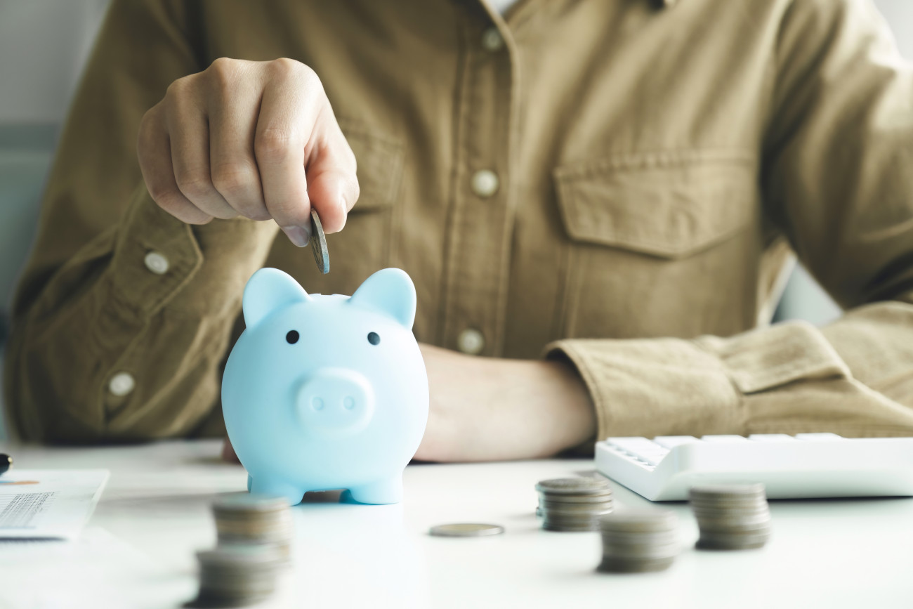 Oseba v rjavi srajci vstavlja kovanec v modrega hranilnega pujska obdan z zloženimi kovanci in kalkulatorjem na mizi