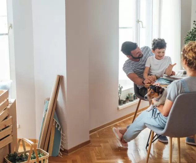 Družina sedi ob velikem oknu v svetlem stanovanju oče se igra s sinom mama pa drži psa v naročju 