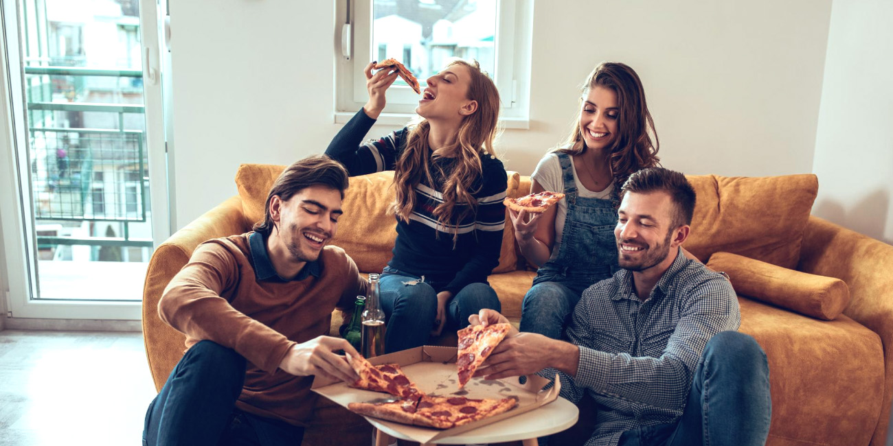 Skupina mladih v dnevni sobi na kavču, v rokah imajo kose pice