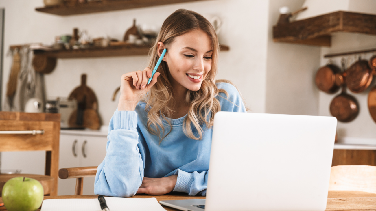 Nasmejana ženska v modri jopici sedi v kuhinji pred prenosnikom in drži pisalo medtem ko se uči ali dela