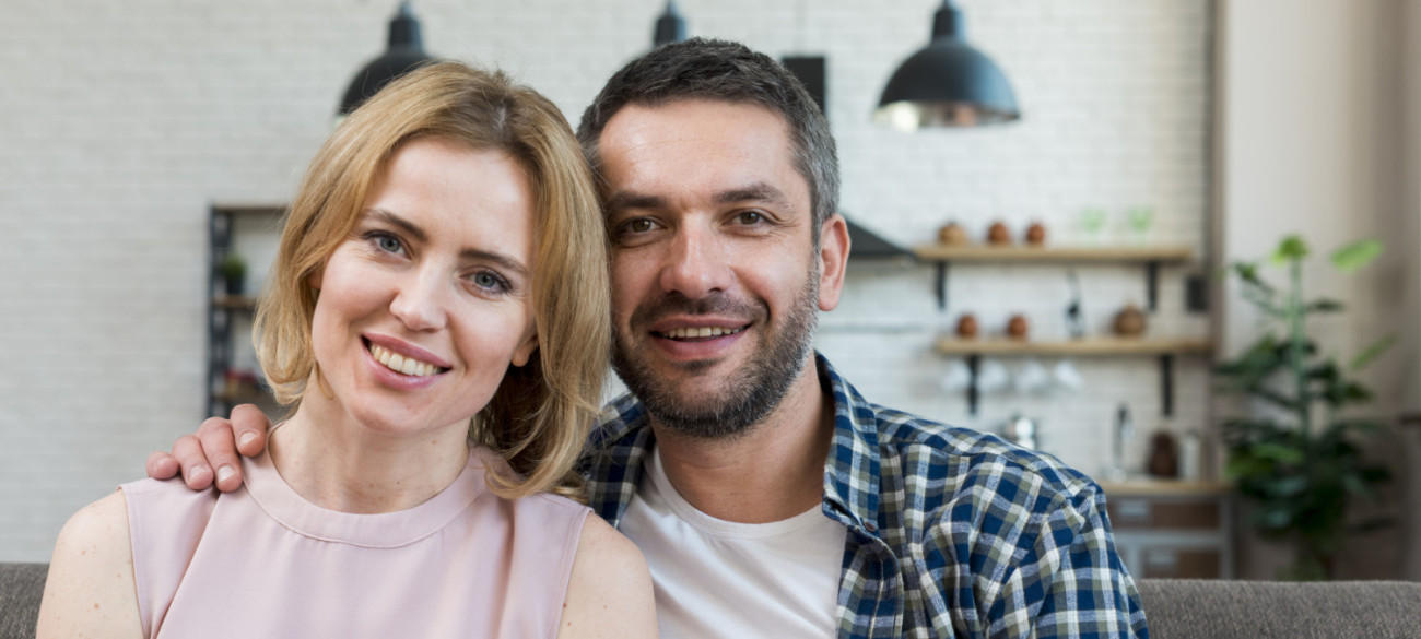 Nasmejan par pozira skupaj na kavču v svetli sodobni kuhinji z belim zidom in visečimi lučmi v ozadju